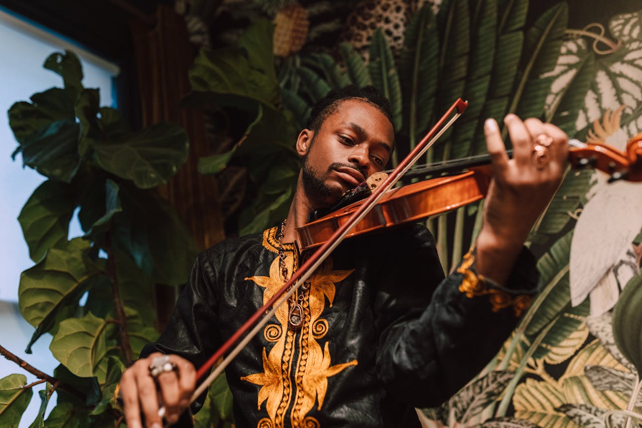 African American man playing violin, wearing traditional clothing, surrounded by indoor plants.
